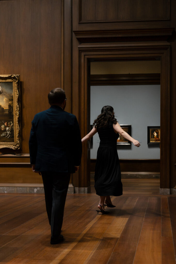 Couple walking away from the camera inside a warmly lit museum gallery filled with framed paintings and display cases.