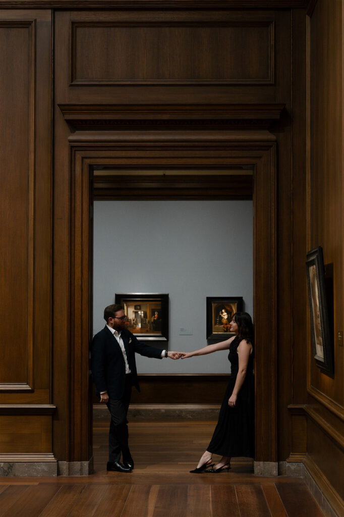 Couple holding hands and facing each other in a museum gallery framed by large classical doorways.