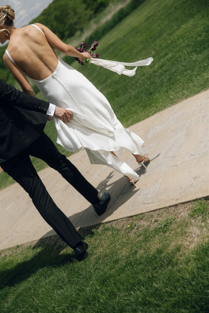 Bride and groom walking together across a paved path in bright midday sun, captured mid-motion from behind as the bride’s dress and bouquet move naturally, showing how direct sunlight adds energy and realism to documentary wedding photography.