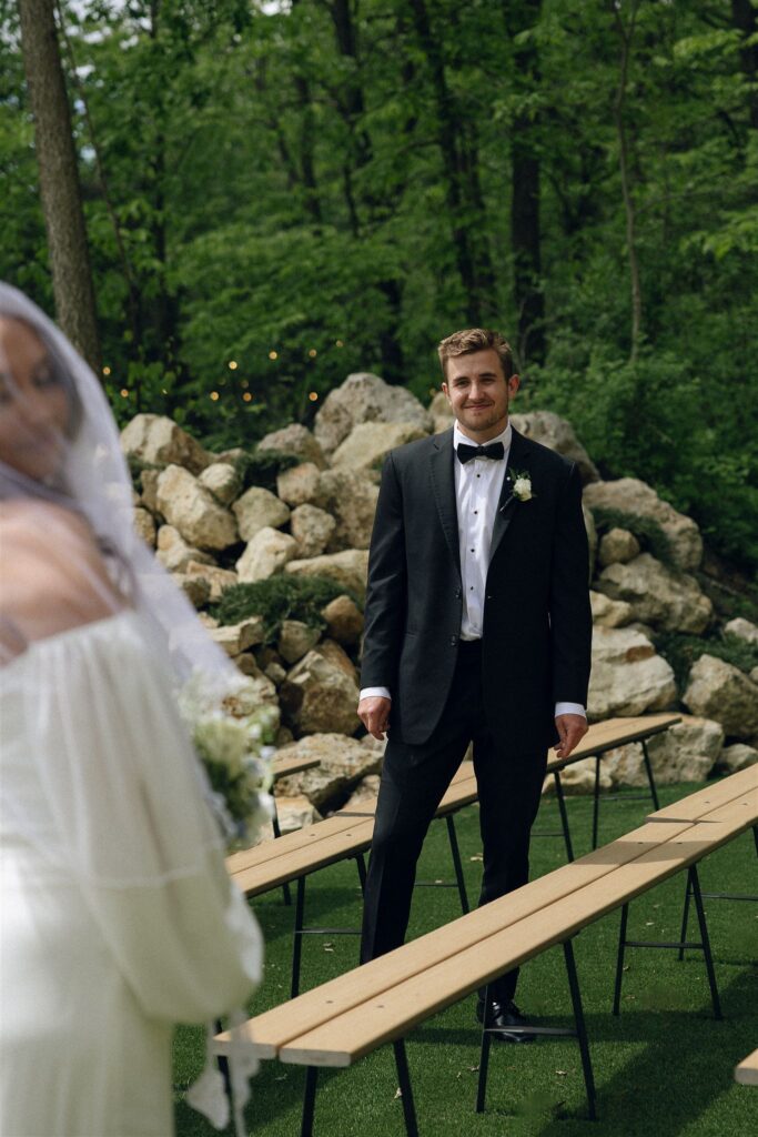 Groom standing at the end of ceremony benches in open shade as the bride approaches from behind, natural diffused light creating a balanced, emotional moment without harsh shadows in outdoor wedding photography.