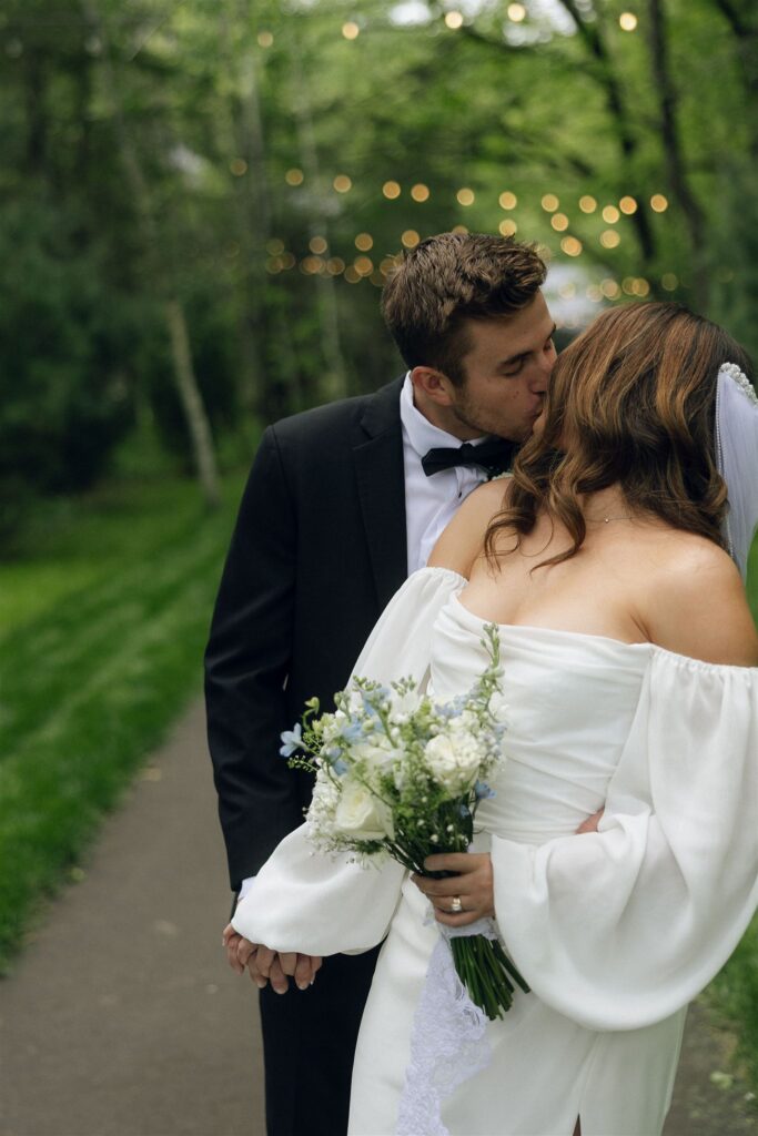 Bride and groom kissing along a tree-lined path in open shade with string lights softly blurred in the background, even lighting preserving the intimate, romantic mood of the moment in wedding lighting photography.