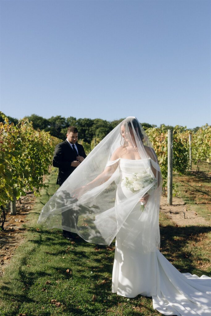 Bride walking through a vineyard in direct sun with her veil lifted by the breeze, sunlight filtering through the fabric to create glow and movement, highlighting how natural light shapes mood in outdoor wedding photography.