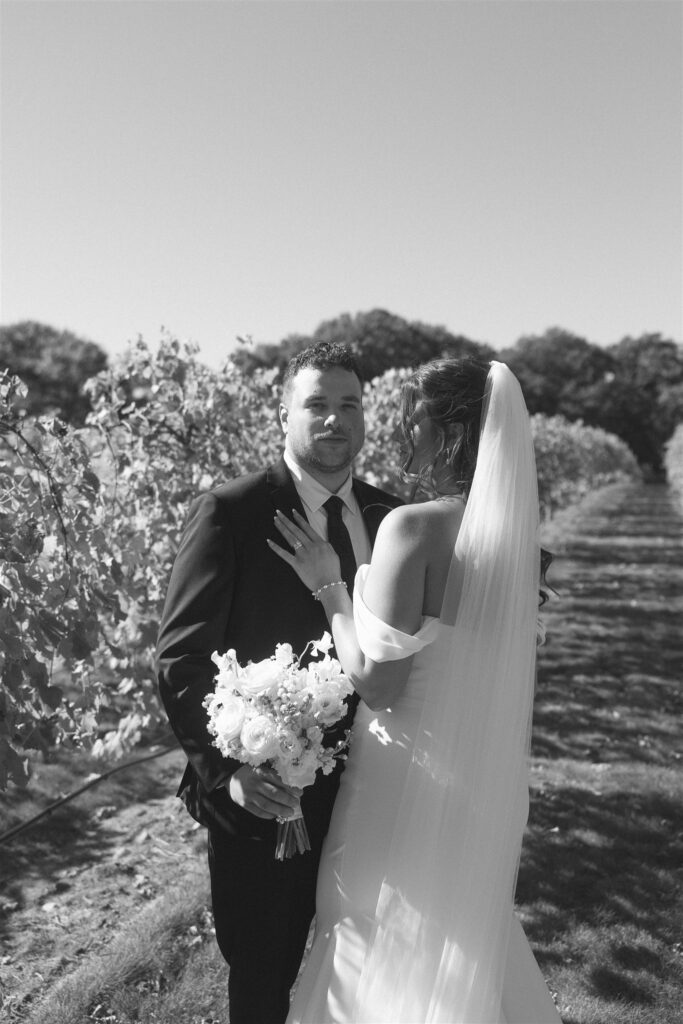 Black-and-white portrait of bride and groom standing between vineyard rows, soft natural light defining their faces and veil without harsh shadows, emphasizing emotion and texture through intentional wedding lighting photography.