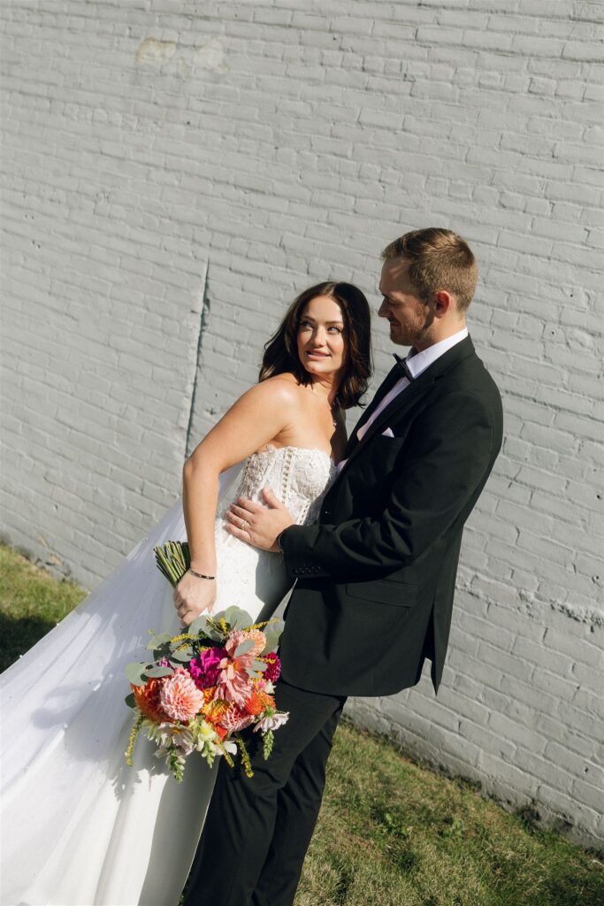 Bride and groom standing close against a white brick wall in strong directional sunlight, crisp shadows and highlights adding contrast and depth, demonstrating how bold light can create drama in wedding venue lighting.