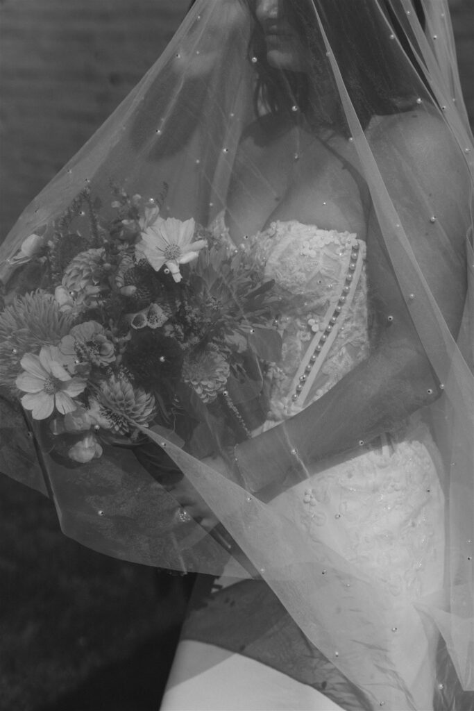 Black-and-white close-up of bride holding her bouquet beneath a beaded veil, soft light filtering through the fabric to create intimacy and quiet emotion, showing how light influences the feeling of wedding photos.