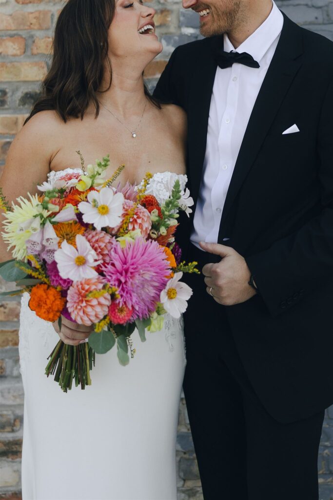 Bride and groom laughing together in open shade against a brick wall, bride holding a colorful dahlia bouquet as soft, indirect light evenly illuminates skin tones and textures in wedding lighting photography.