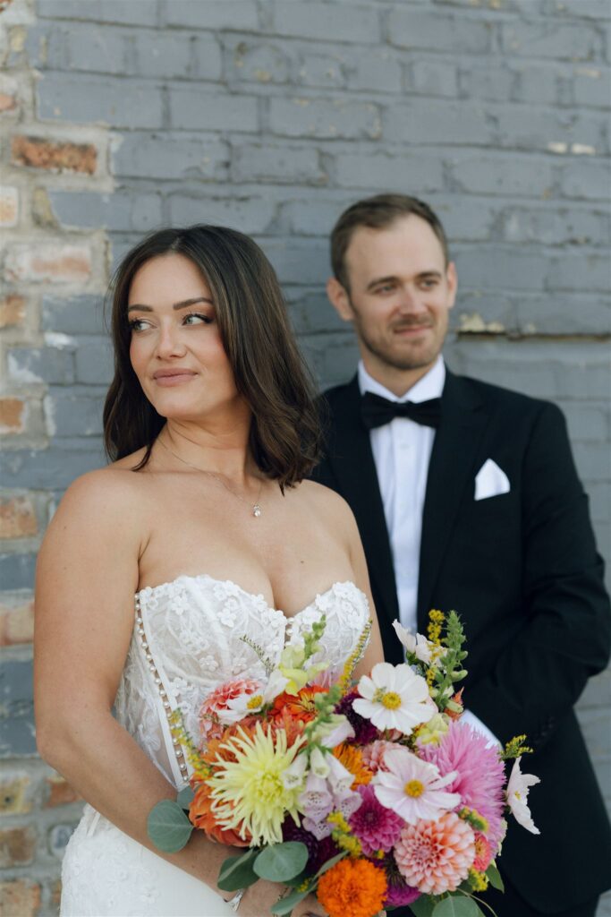 Bride standing in open shade holding a vibrant floral bouquet while the groom stands just behind her, diffused light from the surrounding brick creating soft contrast and a calm, intimate feel in wedding venue lighting.