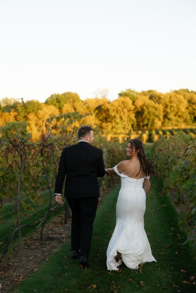 Bride and groom walking hand in hand through vineyard rows during golden hour, warm sunset light casting long shadows and creating a romantic wedding lighting photography moment.