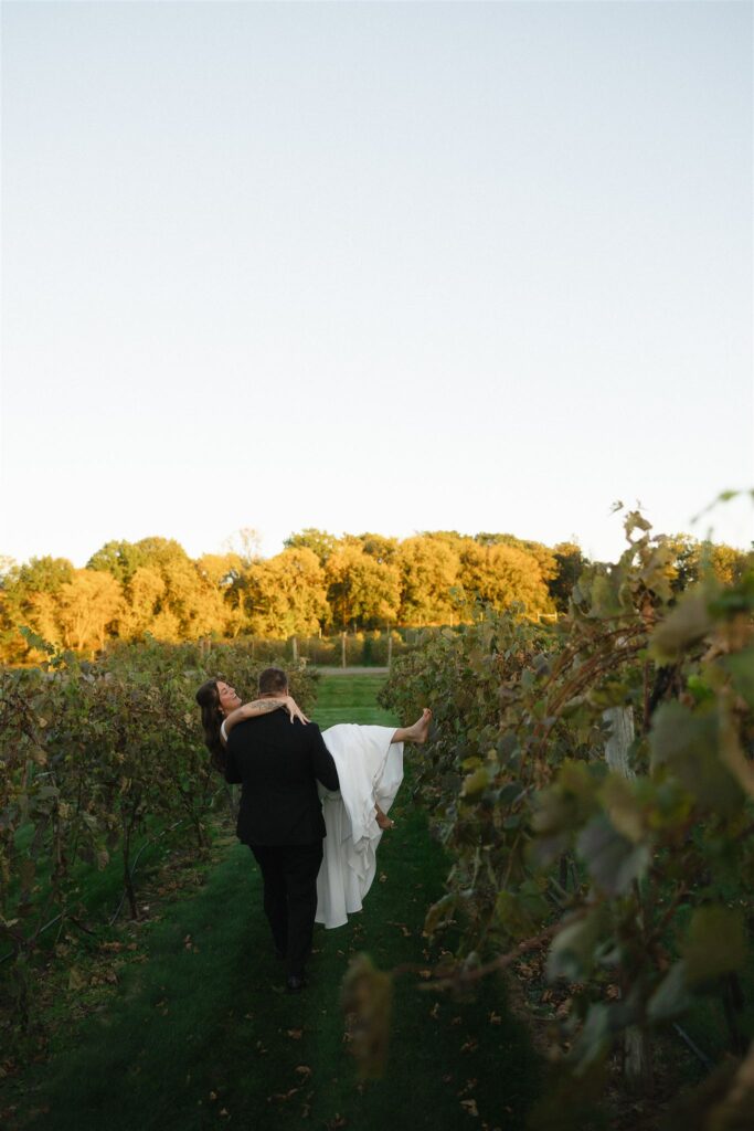 Groom carrying bride through a vineyard at sunset, surrounded by glowing foliage and soft golden hour light that adds warmth and depth to the wedding photos.