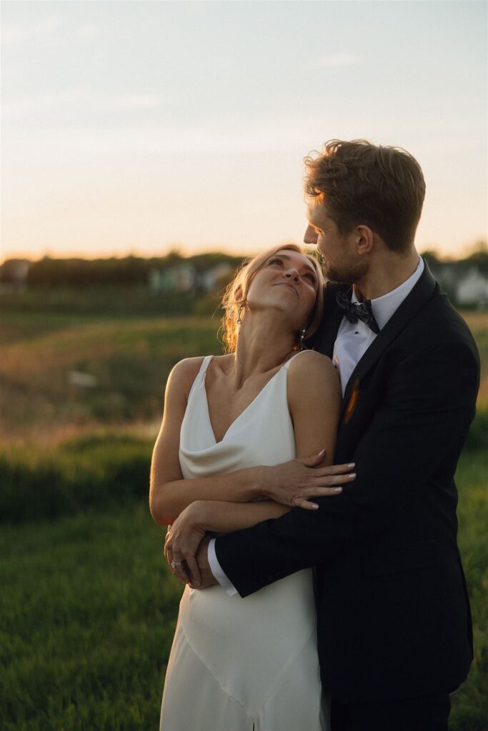 Bride and groom embracing in an open field at golden hour, sun setting behind them and creating soft backlighting that shapes the mood of the wedding lighting photography.