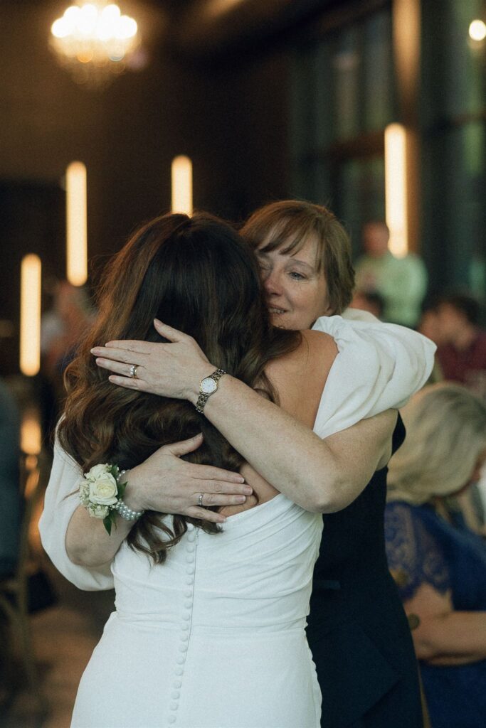 Emotional hug between a bride and her mother during the wedding reception, captured in low-light ambient lighting with warm tones and soft background glow.