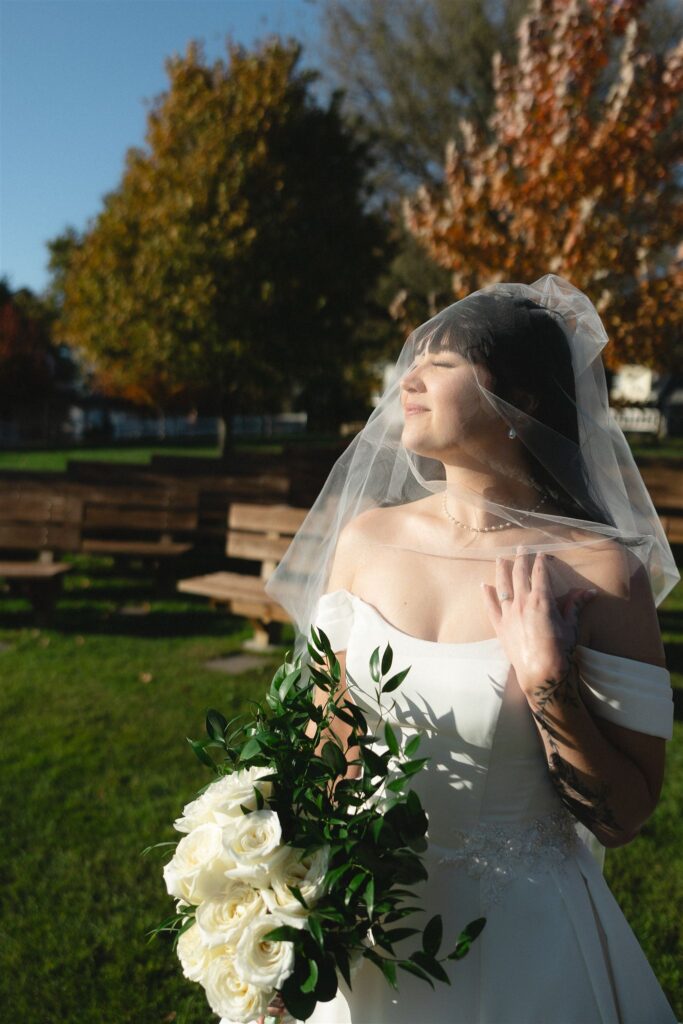 Bride standing outdoors in direct sunlight, eyes closed beneath a sheer veil, holding a white rose bouquet as strong sun creates defined shadows across her dress and arms, demonstrating bold contrast and dimension in wedding lighting photography.