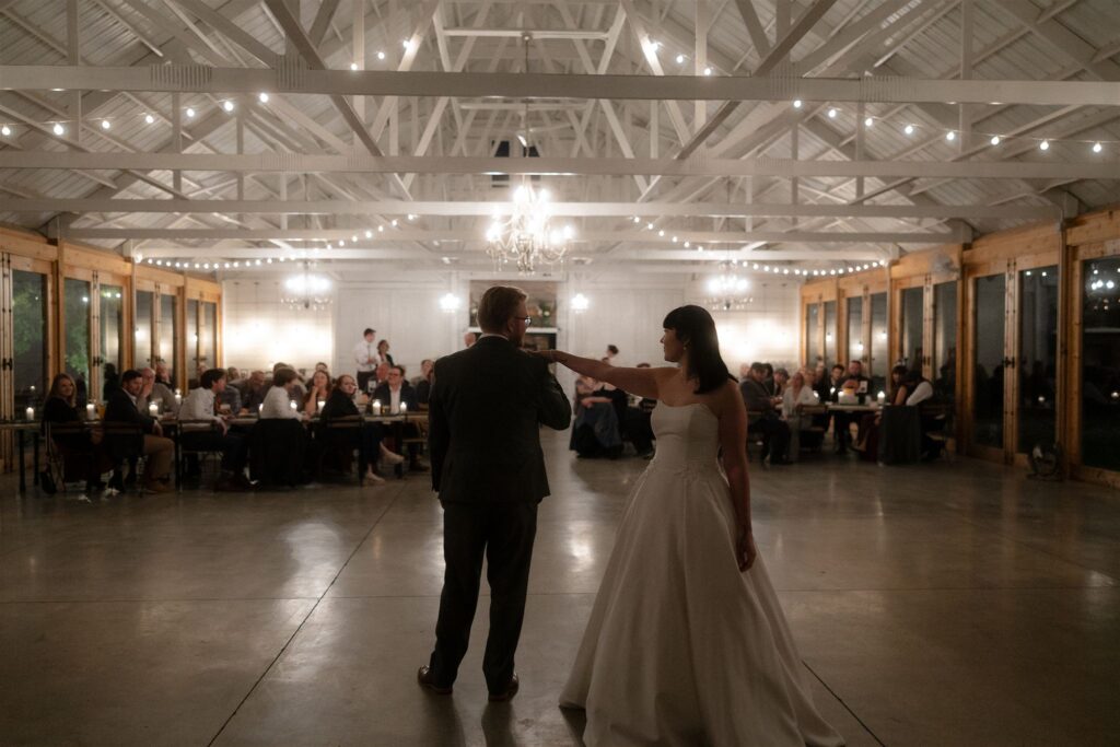 Bride and groom share their first dance in a wedding venue illuminated by chandeliers and string lights, an example of ambient wedding venue lighting captured with natural, emotional wedding lighting photography.